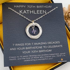 May include: A silver necklace with seven interlocking rings and a purple gemstone pendant. The necklace is displayed on a black card with the text "HAPPY 70TH BIRTHDAY KATHLEEN" and other celebratory text. The card is in a yellow box.