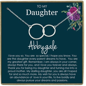 May include: A teal blue card with a silver necklace featuring two interlocking circles. The card has the text "To My Daughter Abbygale" and a message about love and support.