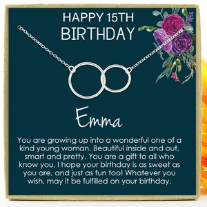 May include: A teal blue card with a silver necklace featuring two interlocking circles. The card has the text "HAPPY 15TH BIRTHDAY Emma" and a message wishing a happy birthday. The card is decorated with a floral design in shades of purple and pink.