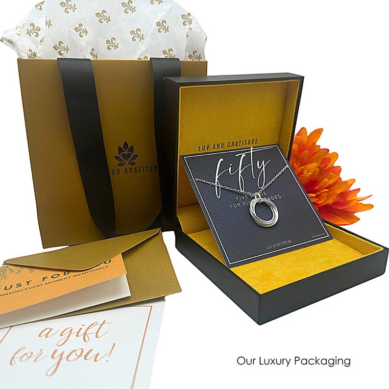 May include: A black gift box with a yellow interior and a card that says "LUV AND GRATITUDE fifty FINE RINGS FOR FIFTY YEARS LUV & GRATITUDE". The box is sitting on a white surface with a brown paper bag and a white tissue paper with a fleur-de-lis pattern.