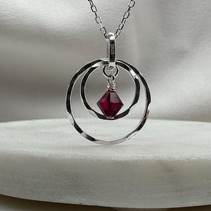 May include: A silver necklace with a pendant featuring two concentric circles and a dangling red crystal.