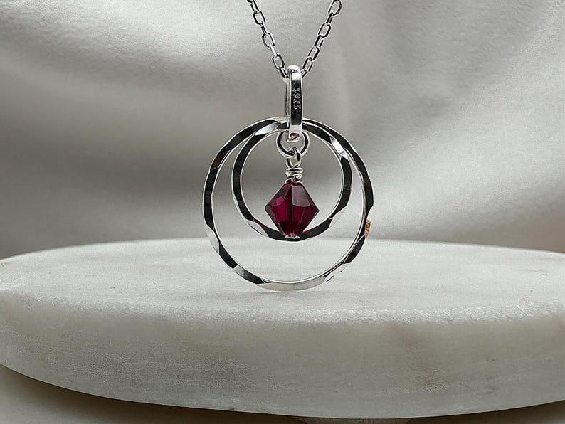 May include: A silver necklace with a pendant featuring two concentric circles and a dangling red crystal.