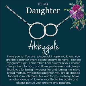 May include: A silver necklace with two interlocking circles hangs on a dark blue background with a floral design. The necklace is inscribed with the name "Abbygale" and the text "To My Daughter". The text below the necklace reads "I love you so. You are so special, I hope you know. You are the daughter every parent dreams to have. You are my greatest gift. Remember, I am always in your corner, always there for you, and I love you forever and always. Thank you for being my daughter and turning me into a proud mother. My darling daughter, you are all I hoped for and so much more. My wish for you is always have an abundance of love in your life, to live boldly and always pursue your dreams and passions."