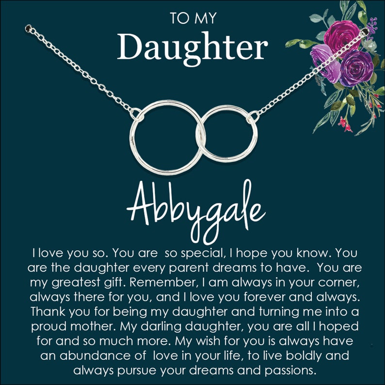 May include: A silver necklace with two interlocking circles hangs on a dark blue background with a floral design. The necklace is inscribed with the name "Abbygale" and the text "To My Daughter". The text below the necklace reads "I love you so. You are so special, I hope you know. You are the daughter every parent dreams to have. You are my greatest gift. Remember, I am always in your corner, always there for you, and I love you forever and always. Thank you for being my daughter and turning me into a proud mother. My darling daughter, you are all I hoped for and so much more. My wish for you is always have an abundance of love in your life, to live boldly and always pursue your dreams and passions."