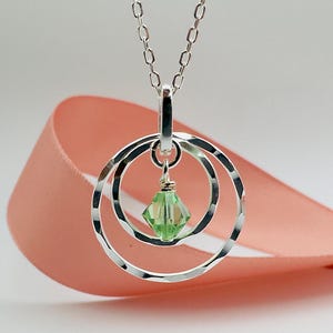 May include: A silver necklace with a pendant featuring three interlocking circles and a light green crystal. The necklace is on a pink ribbon.