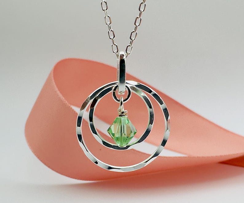 May include: A silver necklace with a pendant featuring three interlocking circles and a light green crystal. The necklace is on a pink ribbon.
