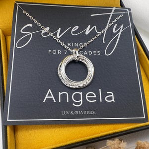 May include: A silver necklace with a pendant of seven interlocking rings, displayed on a black card inside a yellow box. The card reads "Seventy Rings for 7 Decades" and "Angela" with "Luv & Gratitude" below.