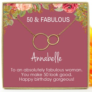 May include: A pink gift box with a gold necklace featuring two interlocking circles. The box has a floral design and the text "50 & FABULOUS" and "Annabelle". The text inside the box reads "To an absolutely fabulous woman. You make 50 look good. Happy birthday gorgeous!"