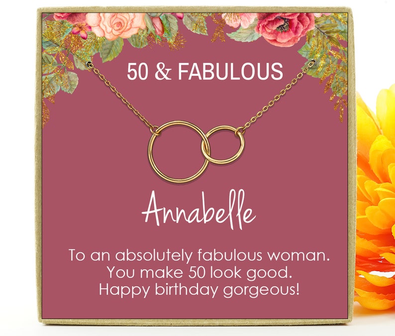 May include: A pink gift box with a gold necklace featuring two interlocking circles. The box has a floral design and the text "50 & FABULOUS" and "Annabelle". The text inside the box reads "To an absolutely fabulous woman. You make 50 look good. Happy birthday gorgeous!"