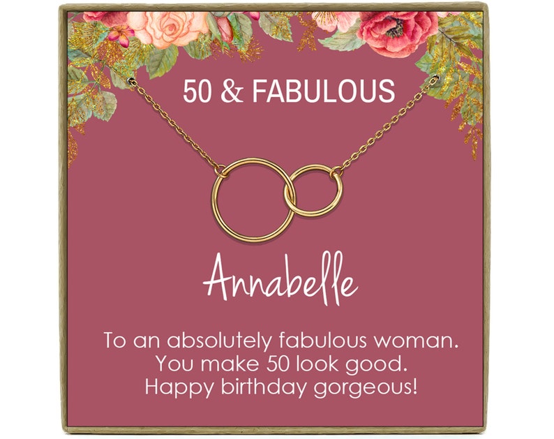 May include: A gold necklace with two interlocking circles, presented in a pink gift box with a floral design. The box has the text "50 & FABULOUS" and "Annabelle" printed on it. The text "To an absolutely fabulous woman. You make 50 look good. Happy birthday gorgeous!" is also printed on the box.