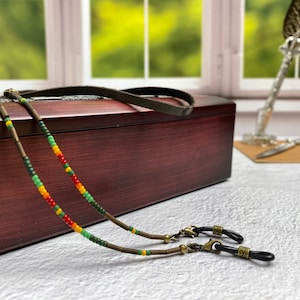 May include: Two beaded eyeglass chains with green, yellow, and red beads. The chains are on a wooden box with a leather strap. The chains have metal clasps at the ends.