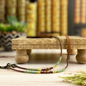 May include: Eyeglass chain with brown, green, and yellow beads. The chain has a brown leather strap and metal connectors. The eyeglass chain is displayed on a small wooden stand with rounded legs, with a blurred background of books.