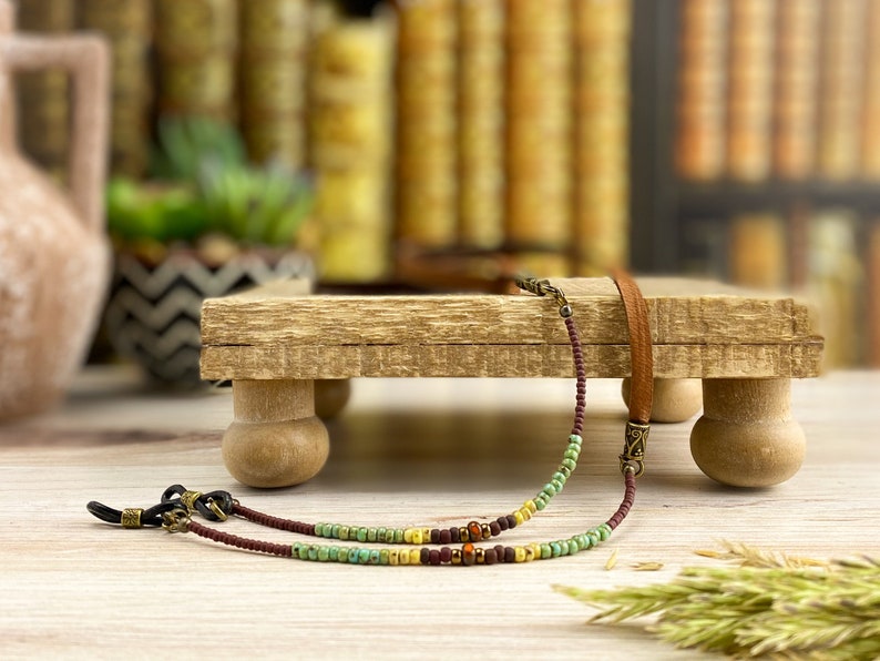 May include: Eyeglass chain with brown, green, and yellow beads. The chain has a brown leather strap and metal connectors. The eyeglass chain is displayed on a small wooden stand with rounded legs, with a blurred background of books.