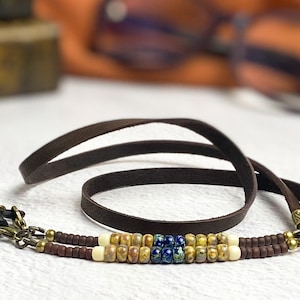 May include: A dark brown leather eyeglass chain featuring a decorative section of small beads in shades of cream, mottled tan, and deep blue. The ends are finished with brass-toned metal findings and black rubber loops for attaching to eyewear frames.
