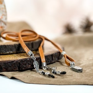 May include: Two brown leather eyeglass chains with silver accents. One chain has a feather charm and the other has a rectangular charm. The chains are displayed on a wooden surface.