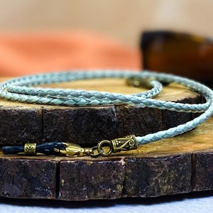 May include: A light blue braided leather eyeglass chain with gold-toned metal clasps. The chain is lying on a wooden surface.