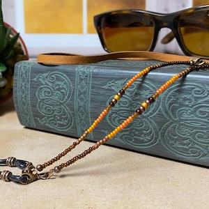May include: Eyeglass chain with a beaded design in shades of brown, orange, and yellow. The chain is draped over a decorative book and near a pair of sunglasses. The chain has metal clasps to attach to glasses.