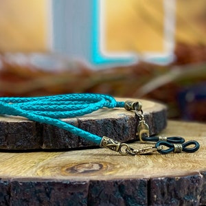May include: A turquoise braided cord with gold-tone metal clasps and black rubber ends. The cord is coiled on a wooden surface.