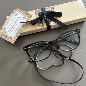 May include: Tortoiseshell eyeglasses with a beaded eyeglass chain and a gift box tied with a black ribbon. The gift box has a feather charm. A tag reads "The Eagle And Hawk Designer Eyeglass Chains and Lanyards".