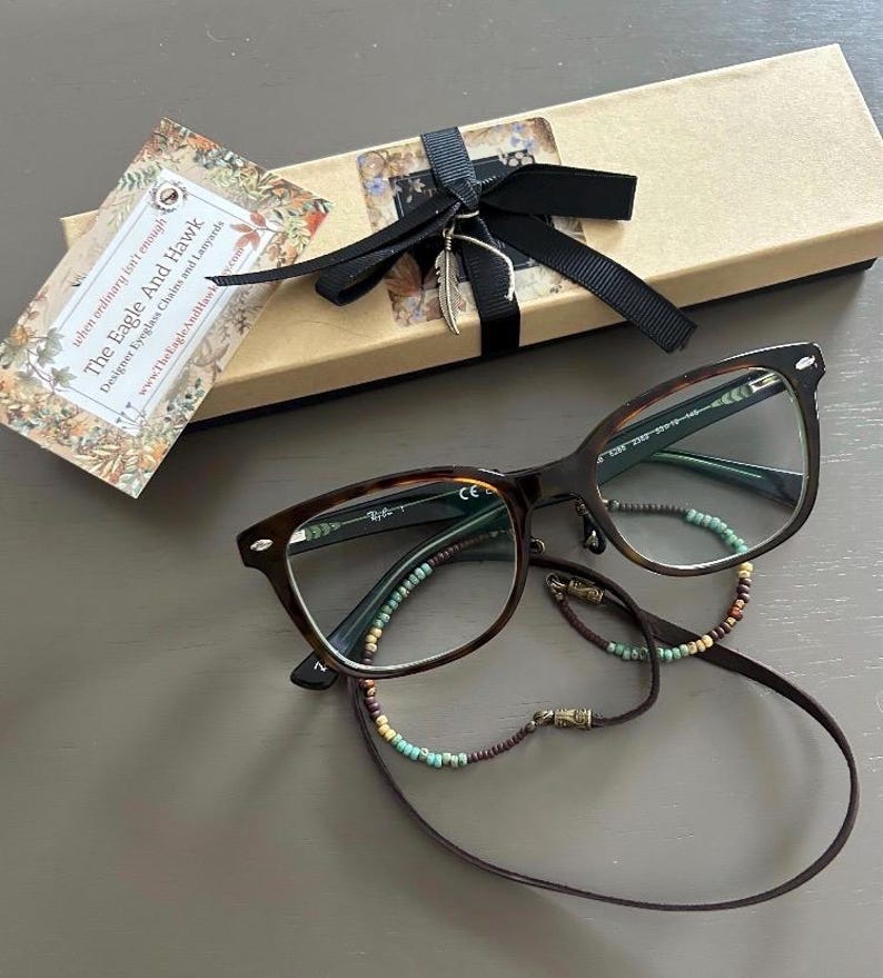 May include: Tortoiseshell eyeglasses with a beaded eyeglass chain and a gift box tied with a black ribbon. The gift box has a feather charm. A tag reads "The Eagle And Hawk Designer Eyeglass Chains and Lanyards".