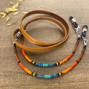 May include: A brown leather eyeglass holder with a beaded chain in shades of orange, blue, and black. The leather strap is coiled, and the beaded chain is attached to the eyeglass holders with silver-colored metal clasps.
