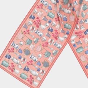 Puede incluir: Una bufanda rosa con un patrón repetido de tazas de té, teteras y flores. La bufanda está hecha de una tela suave y ligera, perfecta para añadir un toque de fantasía a cualquier atuendo.