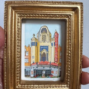 以下が含まれることがあります： オークランド・フォックス・シアターを描いた詳細な水彩画が入った、小さく長方形の装飾的な金色の額縁。このアートワークには、白い文字でOAKLANDと書かれた目立つ縦型の赤い看板がある建物のファサードが描かれています。