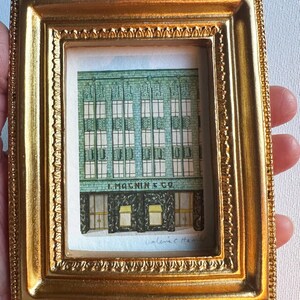 以下が含まれることがあります： 金色の小さな長方形のフレームに、建物のファサードを描いた詳細な水彩画が入っています。この作品は、緑色の建物と、1階の暗い入り口と黄色のオーニングの上に記された「I. MAGNIN & CO.」という文字を描写しています。