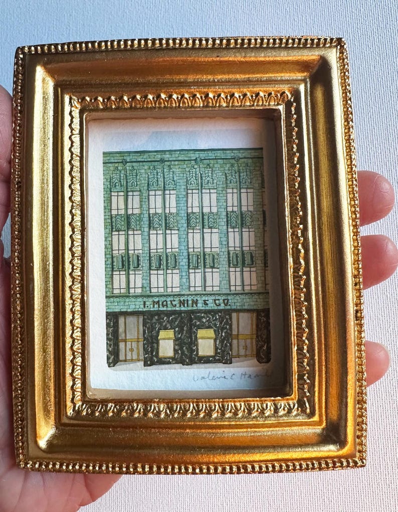 以下が含まれることがあります： 金色の小さな長方形のフレームに、建物のファサードを描いた詳細な水彩画が入っています。この作品は、緑色の建物と、1階の暗い入り口と黄色のオーニングの上に記された「I. MAGNIN & CO.」という文字を描写しています。