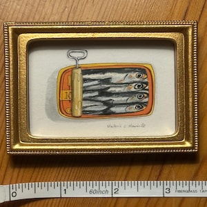 May include: A framed watercolor painting of a sardine can. The can is orange and yellow, with the top pulled back to reveal the sardines. The frame is gold. The painting is signed "Valerie & Marinelle".