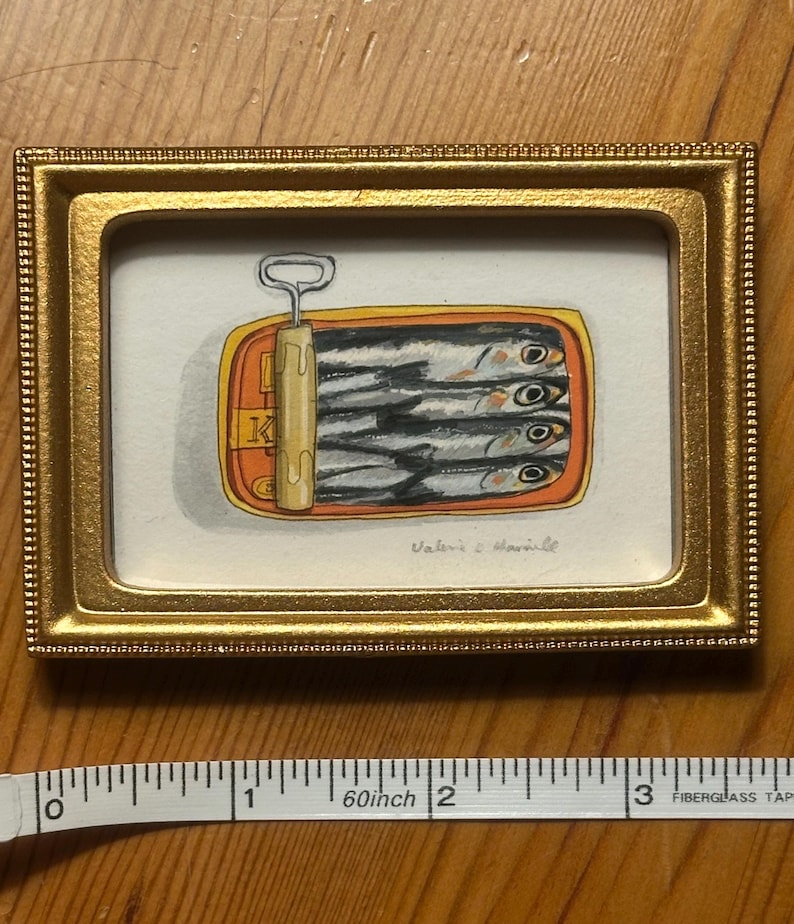 May include: A framed watercolor painting of a sardine can. The can is orange and yellow, with the top pulled back to reveal the sardines. The frame is gold. The painting is signed "Valerie & Marinelle".
