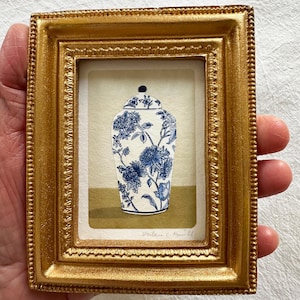 Pode incluir: Uma pequena moldura retangular em tom dourado contendo uma pintura de um pote de gengibre branco com padrões florais azuis. O pote possui uma tampa e assenta sobre uma superfície plana contra um fundo claro. A assinatura é visível abaixo.