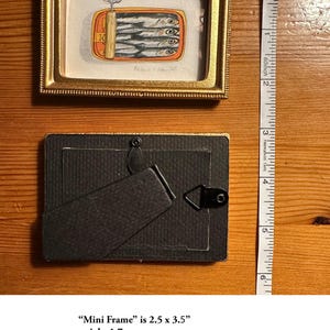 May include: A small, rectangular gold-framed mini frame with a painting of sardines in a can. The frame is 2.5 x 3.5 inches. The back of the frame has a stand. A measuring tape is next to the frame.