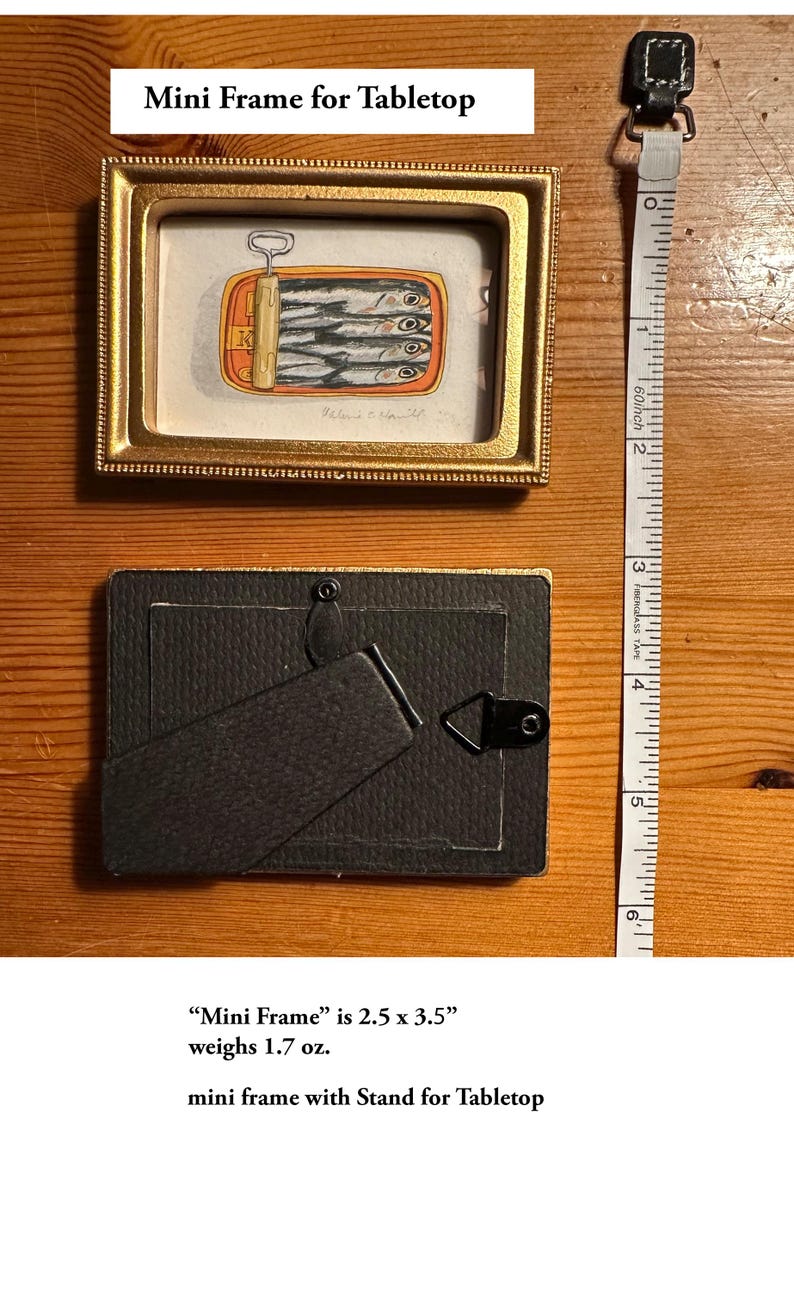 May include: A small, rectangular gold-framed mini frame with a painting of sardines in a can. The frame is 2.5 x 3.5 inches. The back of the frame has a stand. A measuring tape is next to the frame.