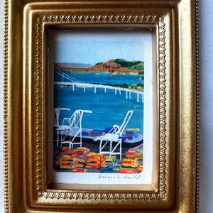 以下が含まれることがあります： 装飾的な金色のフレームに入った小さな長方形の絵画。白いクレーン、カラフルなコンテナ、白い橋、そして遠くに赤い橋が見える港の風景が描かれています。下部にはアーティストのValerie C. Hallの署名が記されています。