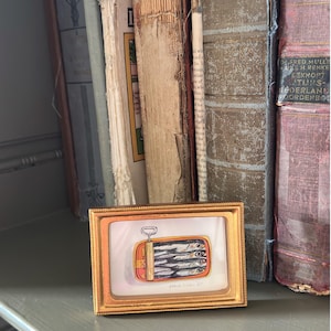 May include: A small framed artwork depicts a can of sardines with a gold frame. The artwork is set against a backdrop of antique books with worn covers. The artwork is on a gray shelf.