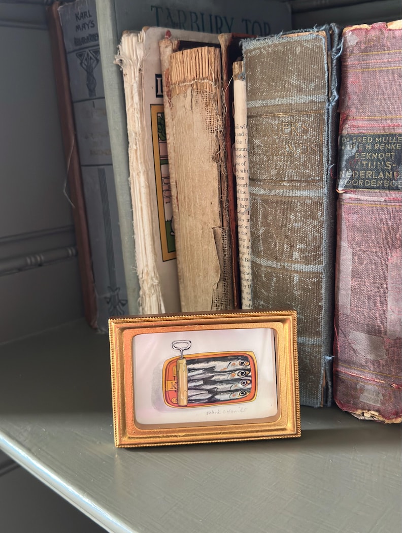 May include: A small framed artwork depicts a can of sardines with a gold frame. The artwork is set against a backdrop of antique books with worn covers. The artwork is on a gray shelf.