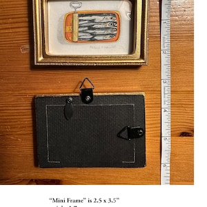 May include: A small, rectangular gold-framed artwork featuring a sardine can illustration. The frame is 2.5 x 3.5 inches and includes a hook for wall hanging. The back of the frame is black. A measuring tape is next to the frame.