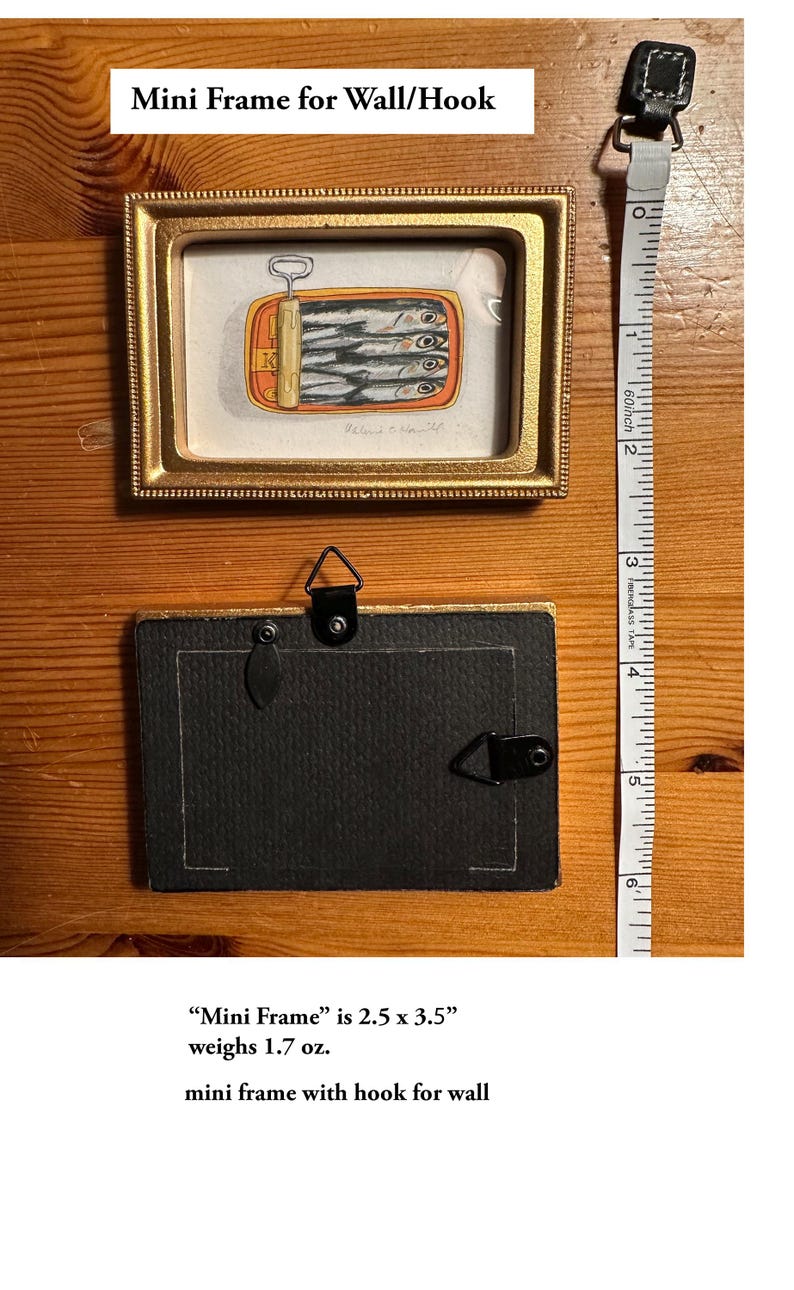 May include: A small, rectangular gold-framed artwork featuring a sardine can illustration. The frame is 2.5 x 3.5 inches and includes a hook for wall hanging. The back of the frame is black. A measuring tape is next to the frame.