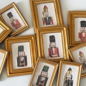 May include: A collection of framed prints featuring illustrations of nutcrackers in various uniforms. The frames are gold-colored with ornate detailing. The nutcrackers are depicted in red, black, and green jackets with tall hats.