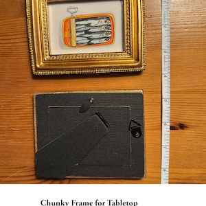 May include: A gold-colored, ornate picture frame with a small painting of sardines in a can. The frame is 4.75 inches by 3.75 inches and includes a stand for tabletop display. The frame weighs 4.3 ounces.