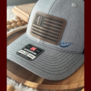 May include: A gray and black trucker hat with a brown leather patch featuring a US flag and the numbers "45 47". The hat has a red label that reads "EST. 1970 RICHARDSON® ORIGINAL 112 TRUCKER ADJUSTABLE".