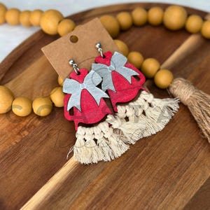 May include: Red wooden earrings with silver bow accents and cream-colored macrame fringe. The earrings are displayed on a brown card and a wooden tray, with wooden beads in the background. A stylish accessory.