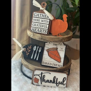 以下が含まれることがあります： 感謝祭をテーマにした装飾が施された、2段の木製トレイ。 「GATHER HERE WITH GRATEFUL HEARTS」のテキストが入った看板、木製の七面鳥、食器のタグ、パイのスライスサイン、そして「Thankful」のサインが含まれています。 秋の装飾。