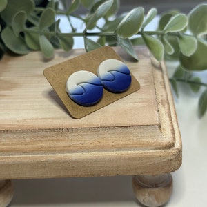May include: A pair of blue and white ombre wave stud earrings on a brown wooden display stand. The stand has the text "Coastal Tides & Co." on the bottom.