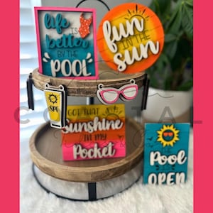 Puede incluir: Un arreglo de bandejas escalonadas con letreros y recortes de madera con temática de verano. Los letreros incluyen frases como "Life is better by the pool" y "Fun in the sun". Otros recortes incluyen gafas de sol, protector solar y un traje de baño. La paleta de colores incluye rosa, turquesa, naranja y amarillo.