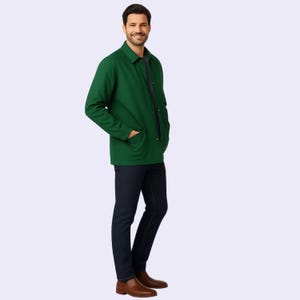 May include: A green jacket with a collared neckline and two front pockets. The jacket is buttoned up and worn with dark blue jeans and brown shoes. The person is smiling.