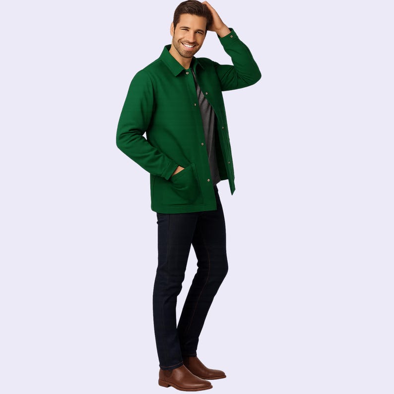 May include: A green jacket with a button-up front and two front pockets. The jacket is paired with a gray shirt, dark blue jeans, and brown boots. The person is smiling and touching their hair.