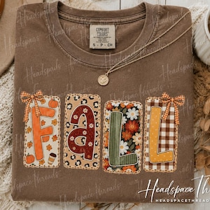May include: A brown crewneck t-shirt featuring the word FALL in large, patterned block letters. Each letter is enclosed in a rectangular patch with distinct designs including pumpkins, leopard print, floral patterns, and brown gingham with bows.