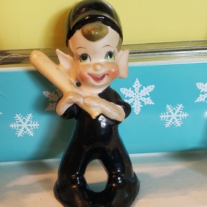 Peut inclure: Une figurine en céramique d'un lutin joueur de baseball portant une casquette noire, un uniforme noir et tenant une batte en bois. Le lutin a un visage souriant avec des yeux verts et des joues roses.